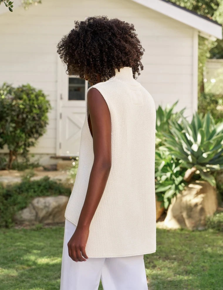 A woman with curly hair wears the FRANK & EILEEN MONTECITO Shell Mock Neck Sweater Shell in Ivory, paired with white pants, standing in a sunny garden with a white cottage and lush greenery in the background.