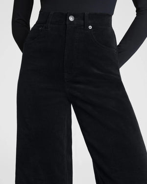 A woman poses against a plain background wearing the SPANX - CORDUROY WIDE LEG JEAN IN BLACK, paired with a fitted black long-sleeve top, black belt, and black heels, her hands in her pockets and expression neutral.