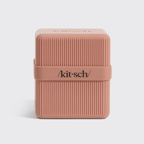 A terracotta, ribbed rectangular container with a matching lid slightly open and leaning against it. The lid features the word “kitsch” in black text. This TSA-approved KITSCH Two Compartment Travel Case is perfect for keeping your beauty essentials organized on the go.