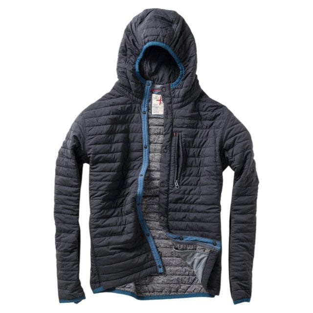 Wearing the RELWEN - WINDZIP HOOD IN BLACK FADE, a black water-resistant jacket with recycled polyester insulation from RELWEN, a person stands on a snowy path in blue pants, covering their face with their hand. Snow falls gently as snow-laden trees fill the background.