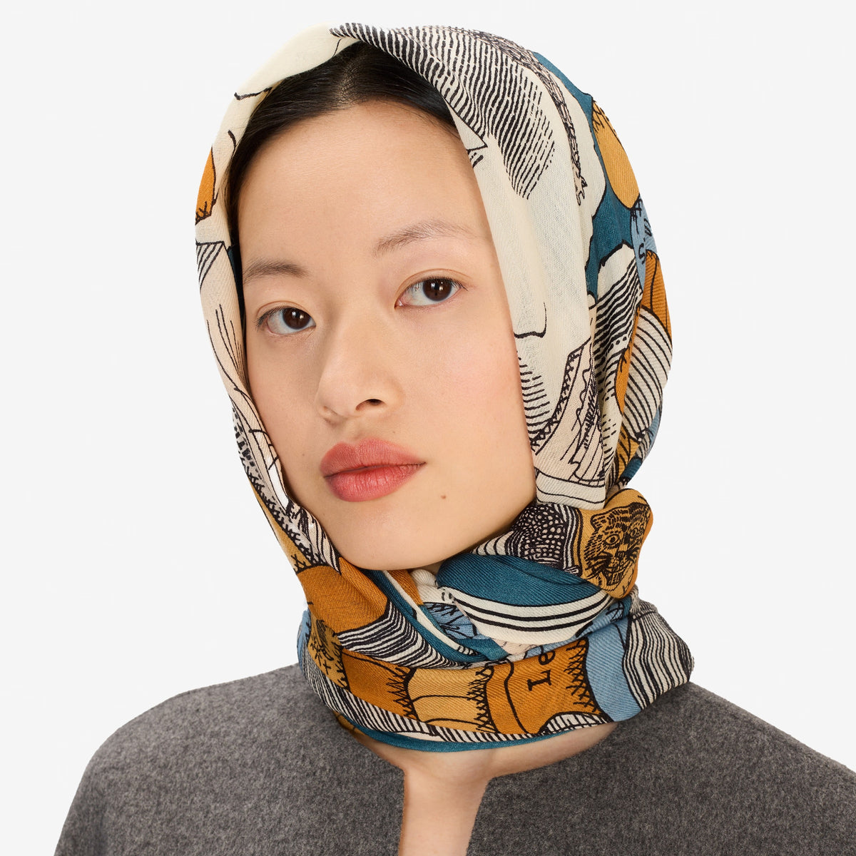 A woman stands facing forward, wearing a grey long-sleeve coat and skirt, black shoes, and the INOUI EDITIONS Stack of Books Wool Scarf in navy, a colorful, hand-printed accessory from INOUI, made in India, on a white background.