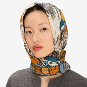 A woman stands facing forward, wearing a grey long-sleeve coat and skirt, black shoes, and the INOUI EDITIONS Stack of Books Wool Scarf in navy, a colorful, hand-printed accessory from INOUI, made in India, on a white background.