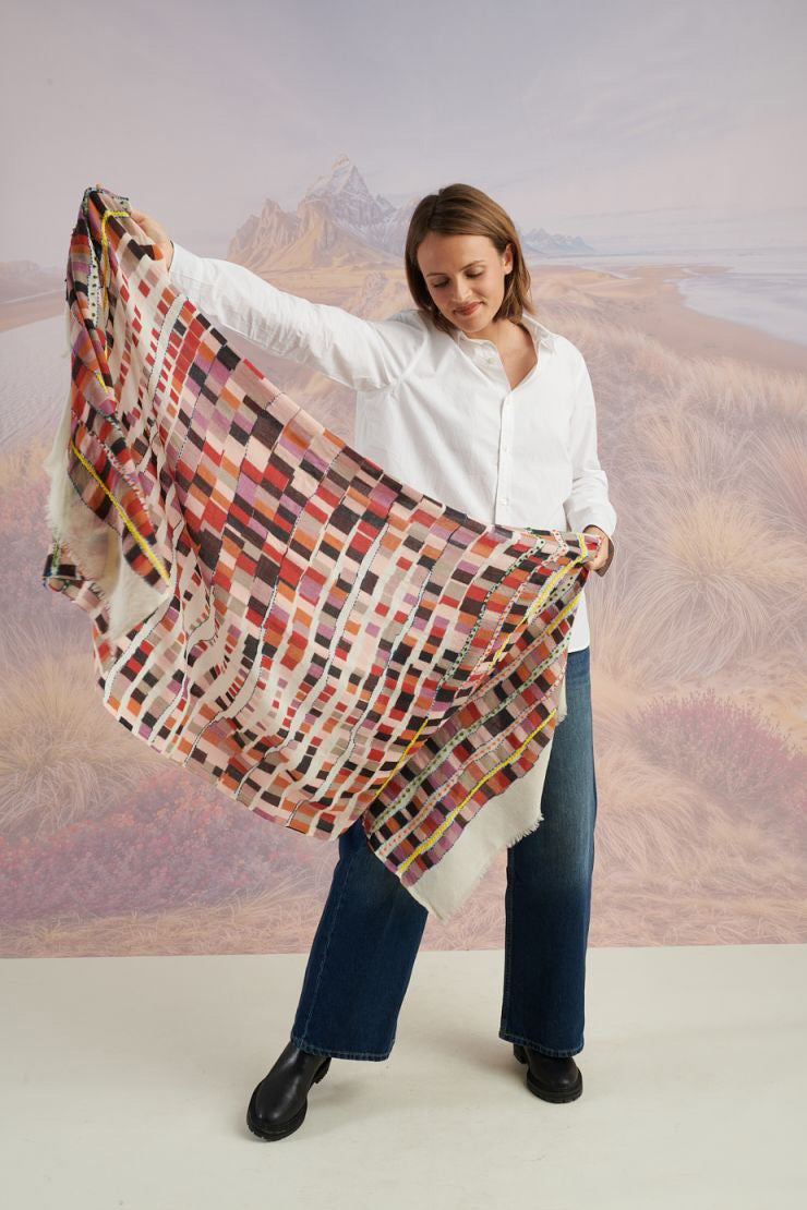 A woman faces the camera, wearing a white button-up shirt, blue jeans, and the STORIATIPIC - ANOUK WOOL SCARF by STORIATIPIC in colorful plaid. The backdrop features soft mountains and grassy plains.