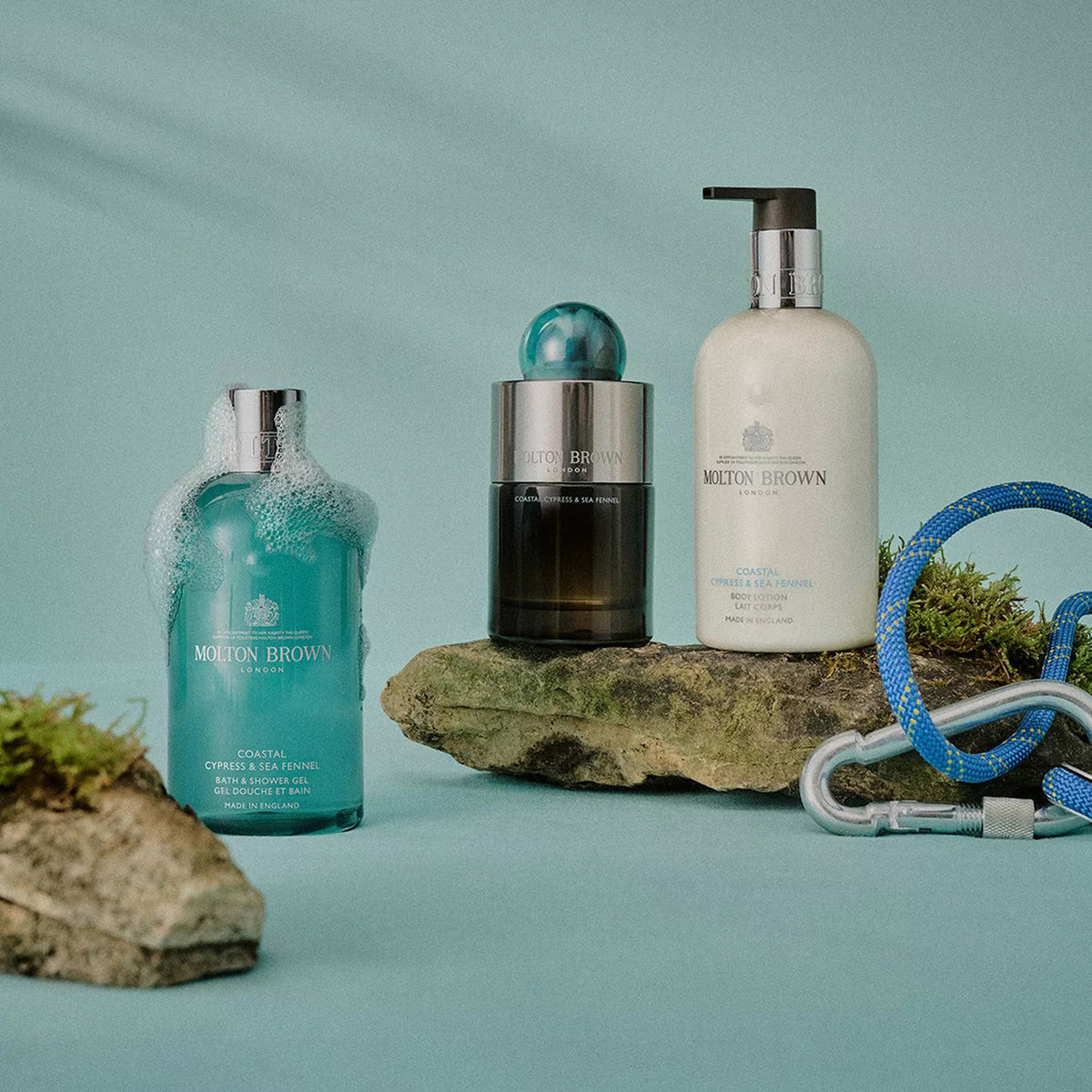 Three Molton Brown bath products, including the Coastal Cypress Body Lotion with a pump, are arranged on mossy rocks against a pale blue backdrop; a climbing rope and carabiners evoke an adventurous, marine-inspired vibe.