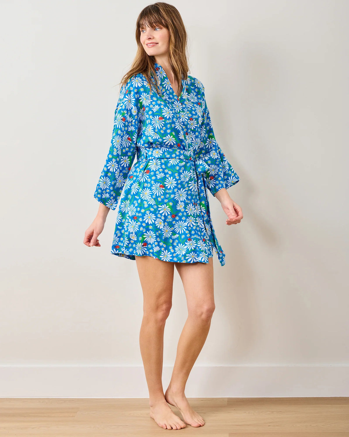 A woman stands barefoot on a wooden floor wearing the PRINTFRESH Satin Mini Robe Lucky Daisy. The blue floral pattern drapes elegantly with long sleeves and a waist tie as she gazes sideways against a light wall, showcasing relaxed elegance and sustainable style.