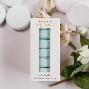 A white box labeled “BAUDELAIRE - PURIFY MINT CLARITY SHOWER STEAMER” contains light blue shower steamers stacked neatly inside, crafted for aromatherapy by BAUDELAIRE. The steamers, infused with essential oil blends, are visible through a clear window, and “Mint Clarity Shower Steamers No. 26” appears at the bottom of the box.