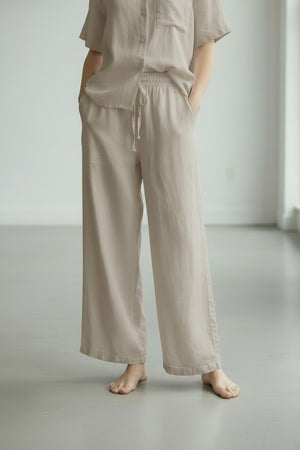 A person wearing the LLEA BLUE - DANA Straight Leg Drawstring Linen Pant in Oatmeal stands barefoot on a light gray floor in a minimal, bright room.