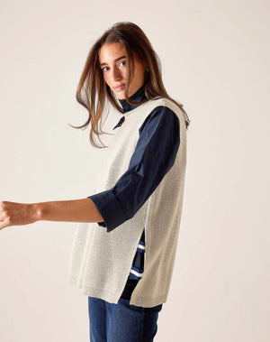 A woman with long brown hair stands sideways against a light background, looking at the camera, wearing the MERSEA - CAMDEN VEST IN DESERT PALM WITH STRIPED TAB by MERSEA over a dark collared shirt and blue jeans.