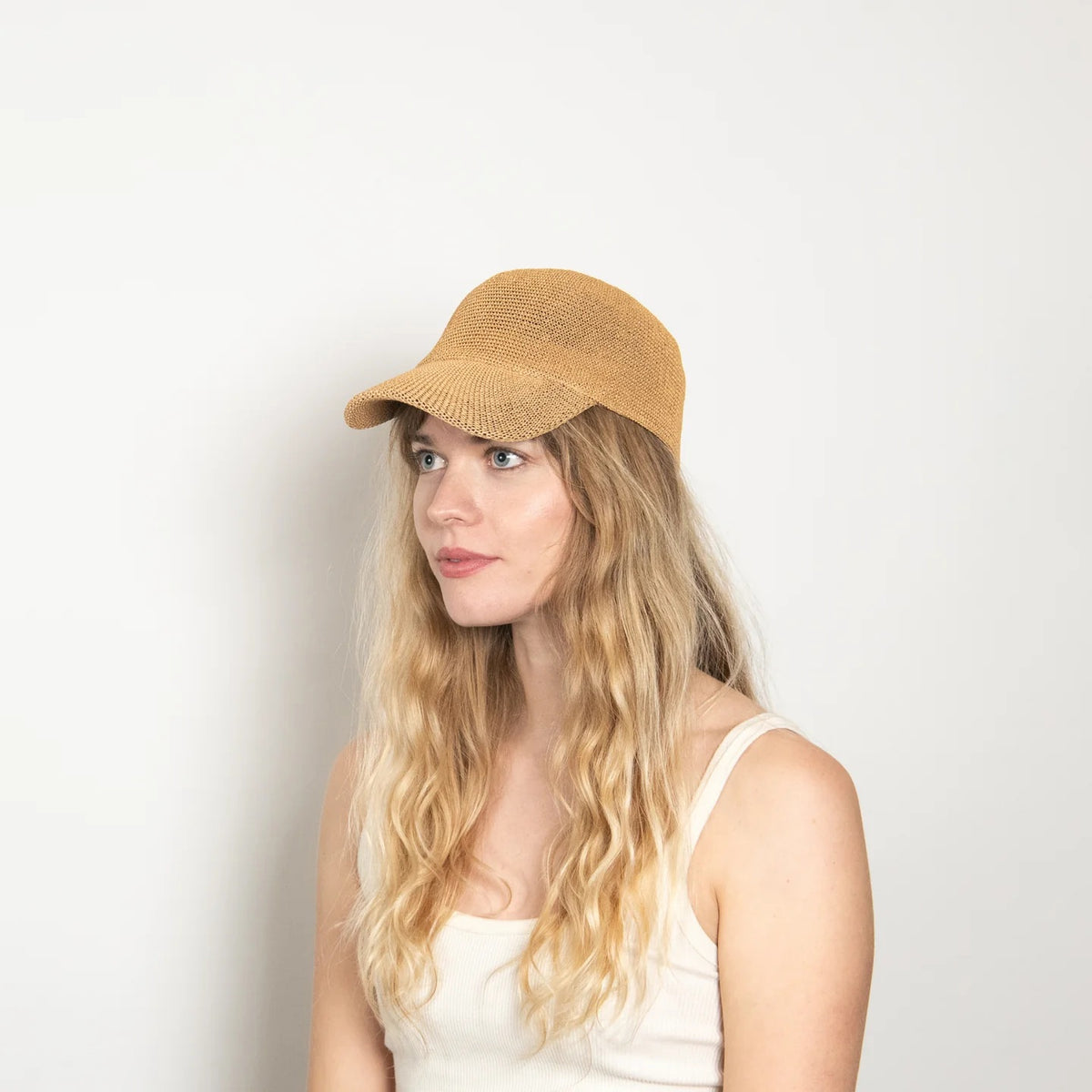 A woman with long wavy blonde hair wears the WYETH HATS - PAULA MESH CAP IN CAMEL by WYETH, featuring UPF 50+ protection, and a white tank top, seated against a plain light-colored background.