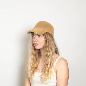 A woman with long wavy blonde hair wears the WYETH HATS - PAULA MESH CAP IN CAMEL by WYETH, featuring UPF 50+ protection, and a white tank top, seated against a plain light-colored background.