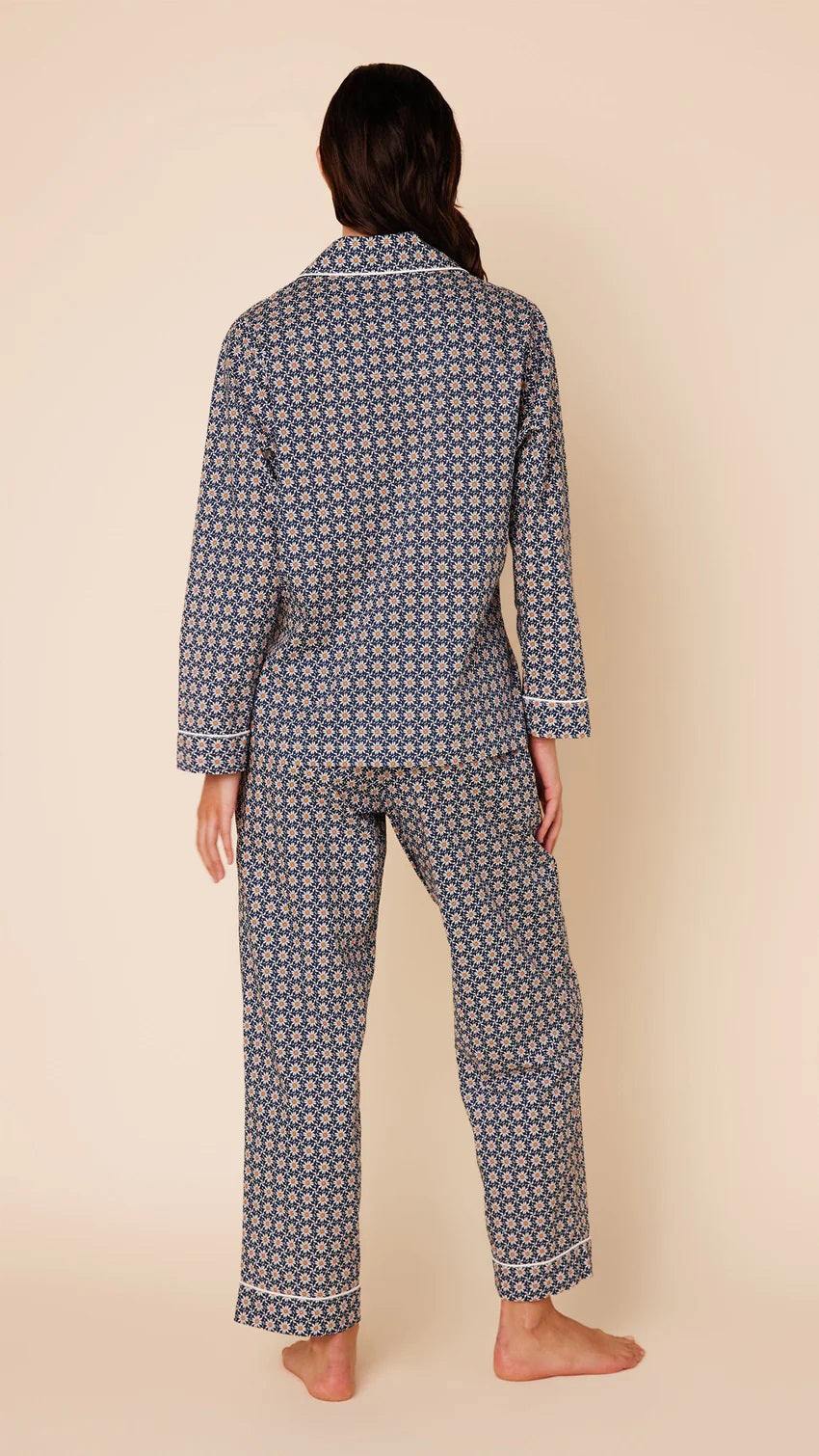 A person stands against a beige background wearing the CAT&#39;S PAJAMAS Newport Daisy Luxe Pima Cotton Pajamas by CAT&#39;S PAJAMAS. The navy blue pajama top features a white geometric pattern, long sleeves, a collar, and white buttons down the front. The matching full-length pajama pants complete the set. The person is barefoot.
