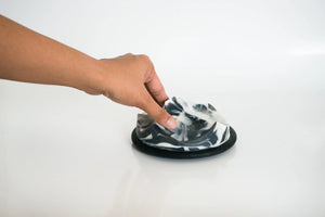 The UME STUDIO UME - JASMINE YLANG YLANG ERODE SOAP, a handcrafted black and white marbled bar shaped like a mountain range, rests on a round black dish against a plain, light background.