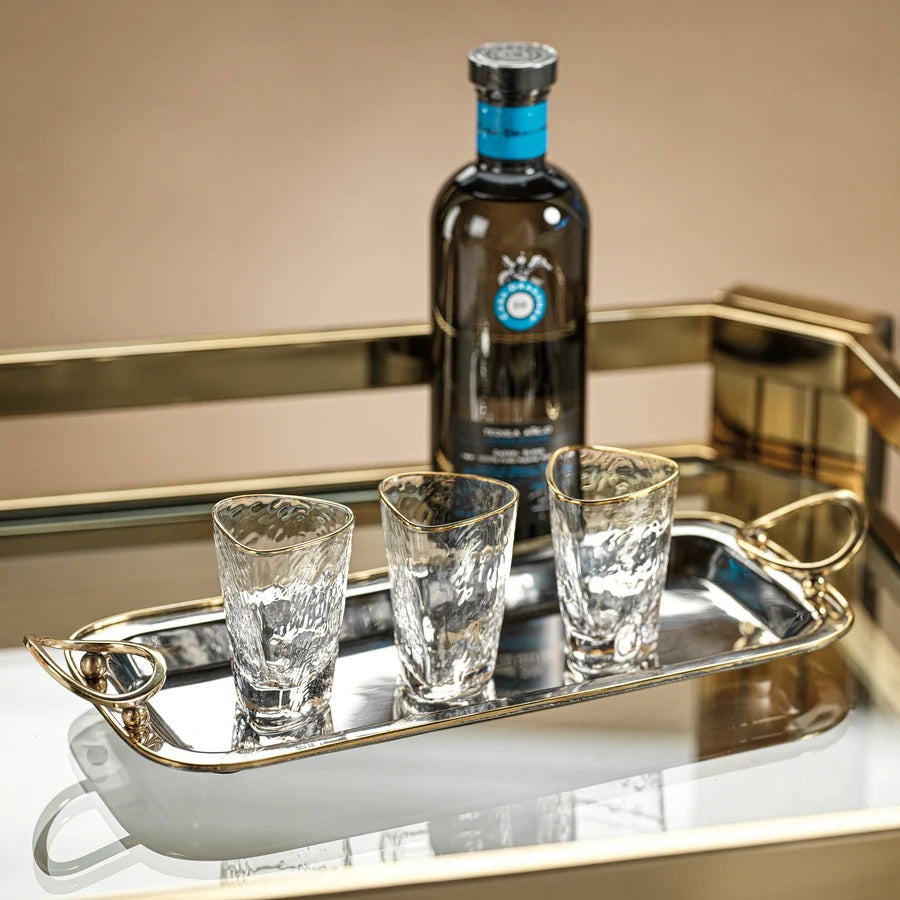 The POLISHED NICKEL AND GOLD TRAY by ZODAX is a rectangular decorative serving tray, crafted from polished nickel with gold trim accents and two circular gold handles, elegantly displayed on a light gray marble surface.