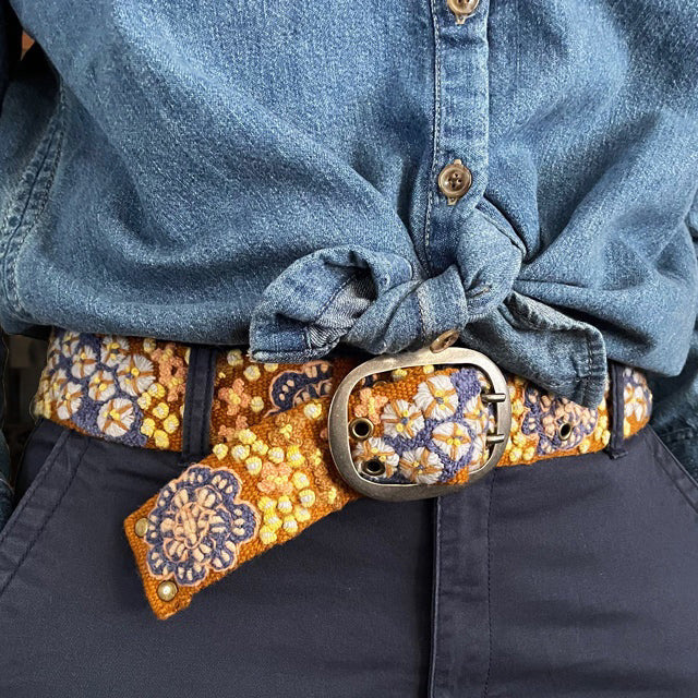 A close-up of a person in a blue denim shirt, dark pants, and the JENNY KRAUSS BLUE SKIES BELT, featuring vibrant floral embroidery and a large oval buckle—handcrafted in Peru.