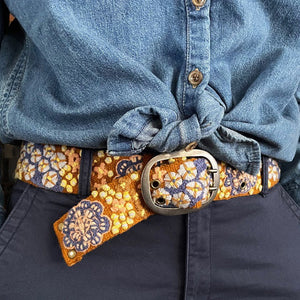 A close-up of a person in a blue denim shirt, dark pants, and the JENNY KRAUSS BLUE SKIES BELT, featuring vibrant floral embroidery and a large oval buckle—handcrafted in Peru.
