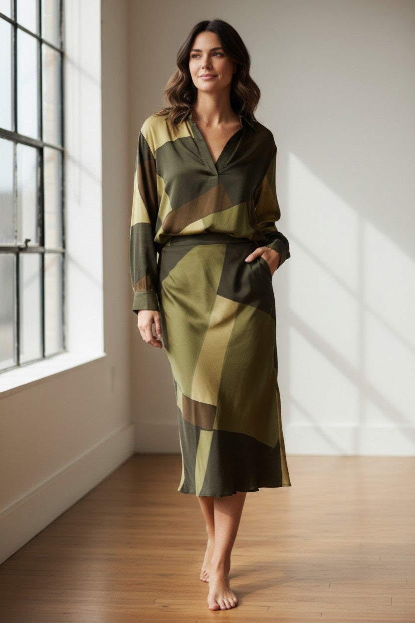 A woman stands in a bright room, smiling. Radiating French elegance, she wears the HARTFORD - CHORUS POPOVER SHIRT IN GREEN by HARTFORD, paired with blue jeans and tan loafers, her hands tucked into her jean pockets. Sunlight pours through large windows beside her.