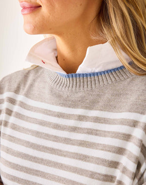 A woman with blonde hair wears the MERSEA - CATALINA MODERN CREWNECK SWEATER in gray and white stripes over a white collared shirt and blue jeans, standing against a plain white wall with a slight smile, one arm bent across her body.