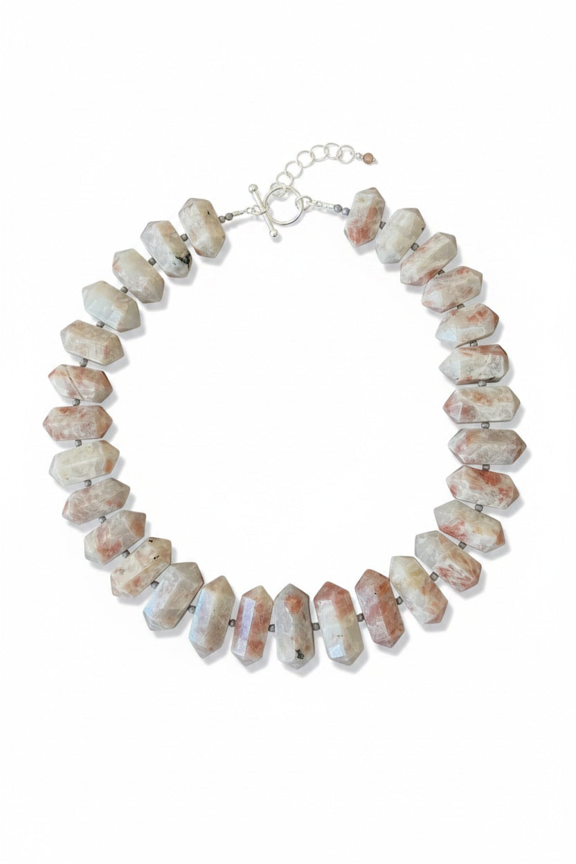 The ANN LIGHTFOOT RAS DU COU OF DOUBLE POINTED SUNSTONE necklace features large, double-pointed sunstone beads in white, gray, and reddish hues with small dark spacers on a white cord.