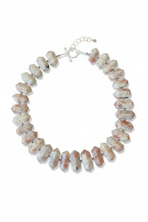 The ANN LIGHTFOOT RAS DU COU OF DOUBLE POINTED SUNSTONE necklace features large, double-pointed sunstone beads in white, gray, and reddish hues with small dark spacers on a white cord.