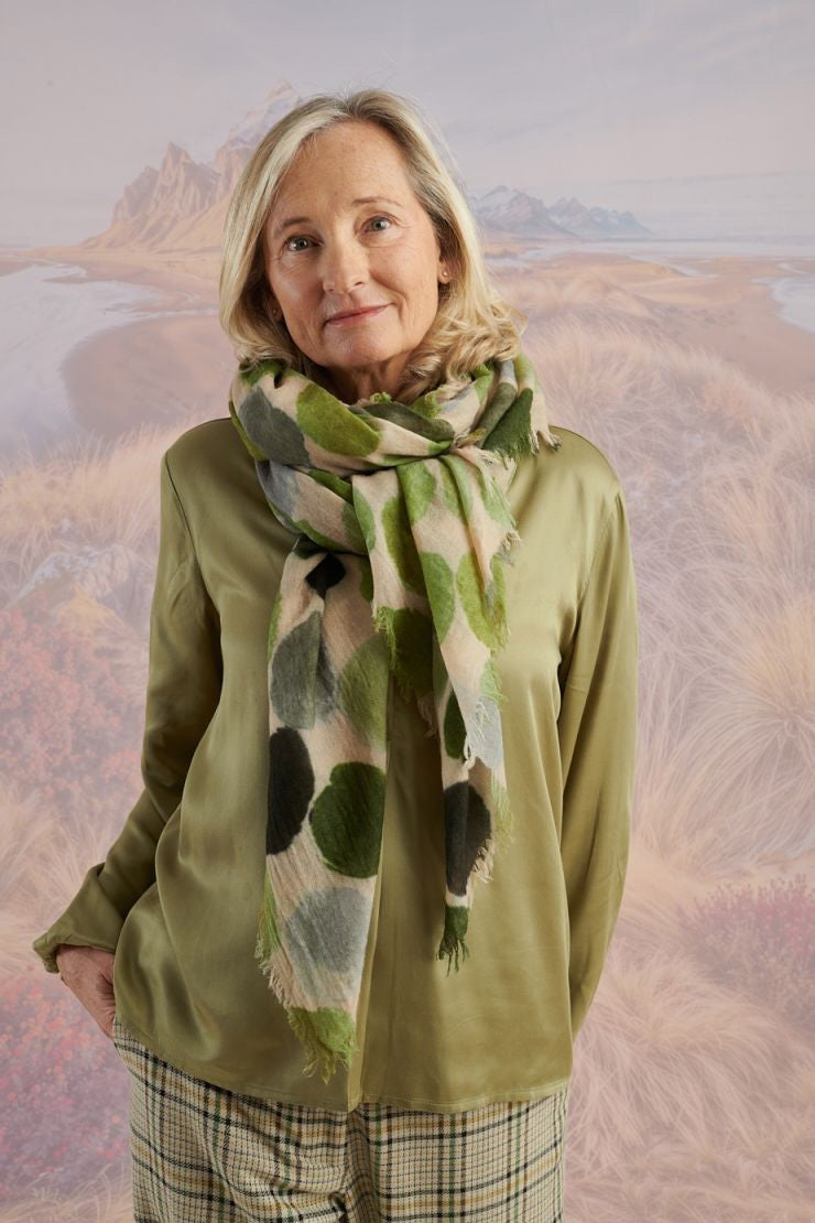 A woman with blonde hair displays the STORIATIPIC - BETTY WOOL SCARF, a large rectangular wool accessory with earth-toned polka dots, set against a scenic mountain landscape.