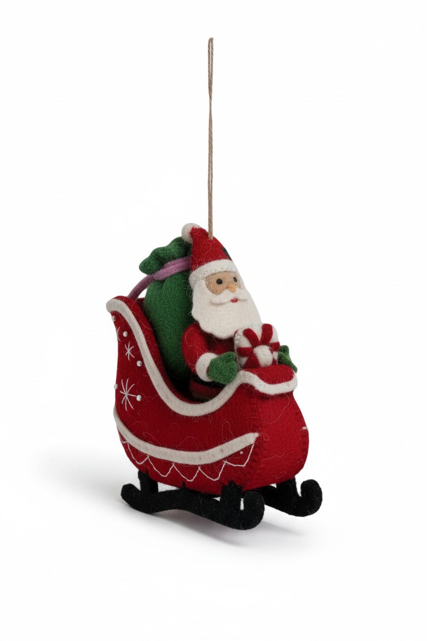A CREATIVE COOP WOOL FELT SANTA IN SLEIGH ORNAMENT featuring a handcrafted Santa Claus sitting in a red sleigh, holding a gift and carrying a green sack.