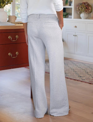 Indoors, a person wears FRANK & EILEEN's RILEY Wide Leg Trouser Sweatpant in light gray with a button waistband and a white long-sleeve top, one hand in their pocket. A red desk, wooden floor, and white cabinets complete the California Heritage Collection scene.