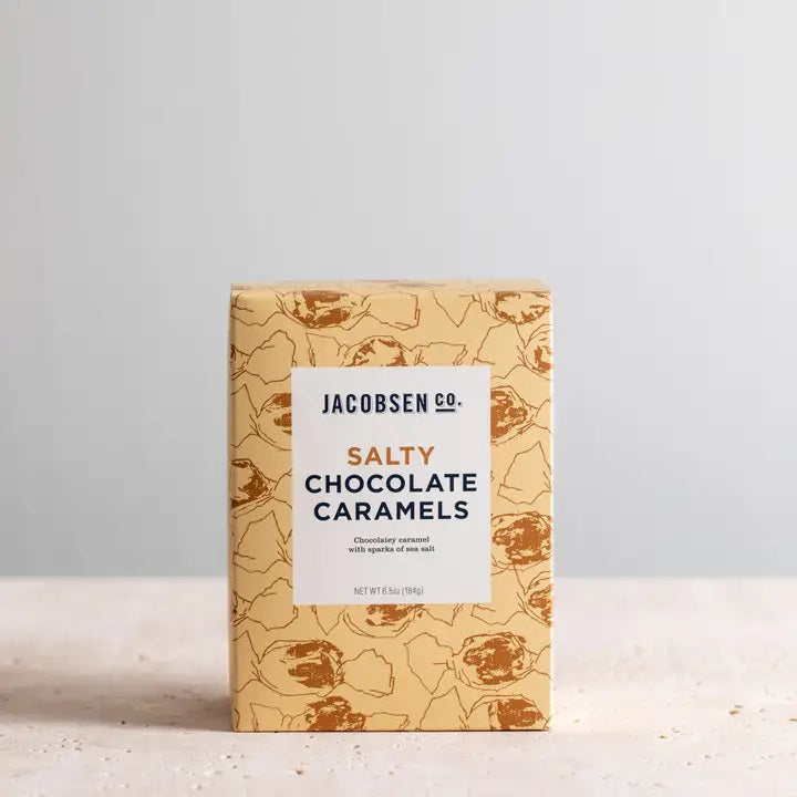 A rectangular box of JACOBSEN SALT CO. CHOCOLATE CARAMELS WITH SEA SALT stands on a light surface, featuring tan packaging with brown abstract patterns and a white label displaying the product name in black and gold.