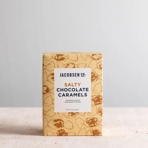 A rectangular box of JACOBSEN SALT CO. CHOCOLATE CARAMELS WITH SEA SALT stands on a light surface, featuring tan packaging with brown abstract patterns and a white label displaying the product name in black and gold.