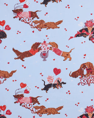 A woman with long, wavy brown hair stands barefoot against a white wall, wearing the PRINTFRESH Dachshund Date Short Sleeve Top & Cropped Pants Pajama Set in Blue. The organic cotton set from PRINTFRESH features short sleeves and cropped pants adorned with a charming dachshund print. She smiles slightly.