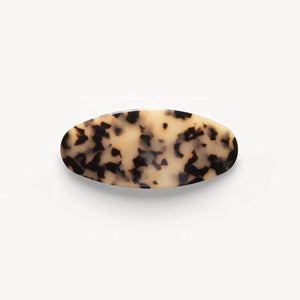 A person with wavy brown hair styled in a low twist, secured by the MACHETE Jumbo Oval Barrette in Blonde Tortoise, wears gold hoop earrings and a sleeveless black top, facing away from the camera against a white background.