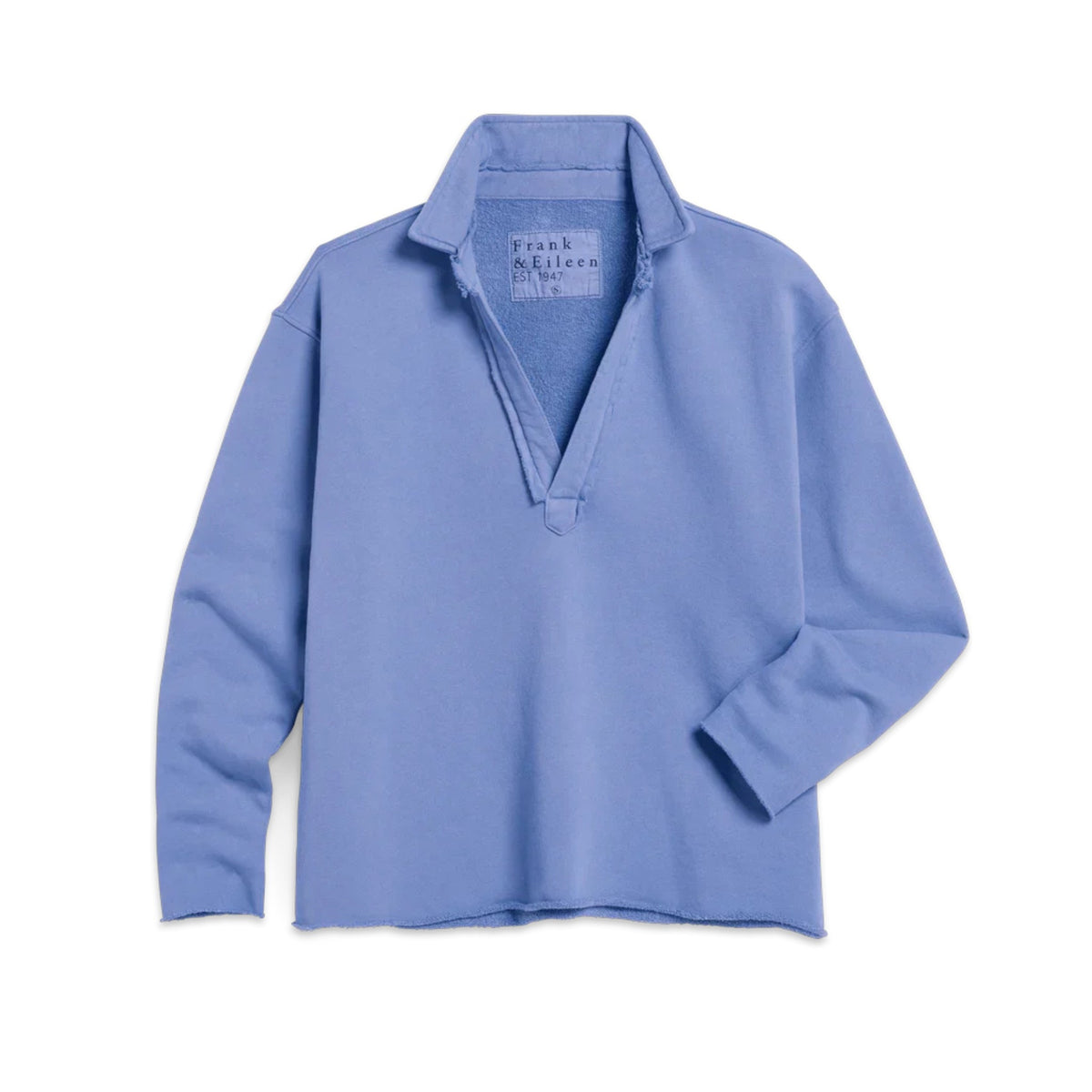 A woman with curly hair smiles while walking outdoors in a FRANK &amp; EILEEN Patrick Popover Henley in Triple Fleece Frost, paired with white pants and gray shoes. She holds a gray jacket in a sunlit, garden-like setting.