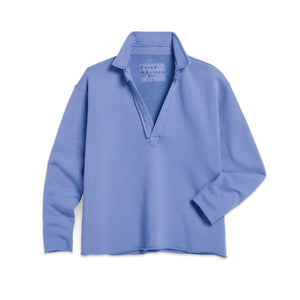 A woman with curly hair smiles while walking outdoors in a FRANK & EILEEN Patrick Popover Henley in Triple Fleece Frost, paired with white pants and gray shoes. She holds a gray jacket in a sunlit, garden-like setting.