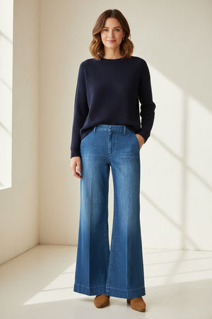 Indoors, someone wears a white long-sleeve shirt, white sandals, and FRANK & EILEEN's WEST VILLAGE NYC WIDE LEG DENIM TROUSER IN 1984 WASH, with their left hand in their pocket. The kitchen background features baskets and wooden cabinets.
