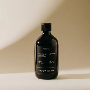 A black bottle of PRESTON - BODY WASH labeled "Hydrating Avocado Body Wash Leather" is centered against a beige background. Made in the USA with plant-based ingredients, this 16 oz (500 ml) moisturizing body wash from the brand PRESTON ensures hydrated skin without harsh chemicals.