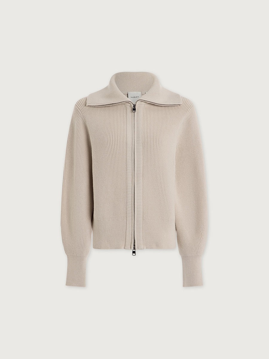 A person wearing a dark brown VARLEY - ENRICO ZIP-THROUGH KNIT JACKET featuring a ribbed soft viscose knit, a high collar, and a front zipper, ideal for cool weather layering.