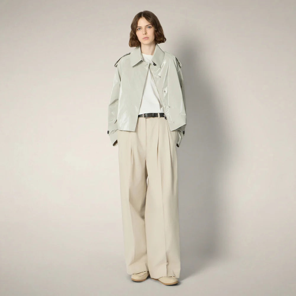 A woman with shoulder-length brown hair wears the SAVE THE DUCK - GAIA JACKET, a shiny light gray cropped trench-style jacket with wide sleeves and large black buttons, paired with high-waisted beige pleated trousers against a plain background.