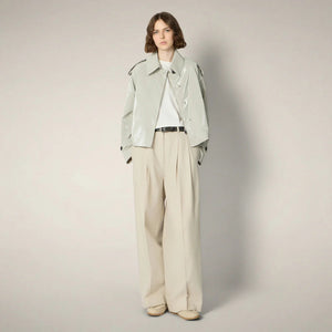 A woman with shoulder-length brown hair wears the SAVE THE DUCK - GAIA JACKET, a shiny light gray cropped trench-style jacket with wide sleeves and large black buttons, paired with high-waisted beige pleated trousers against a plain background.
