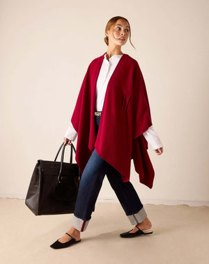 A woman in the MERSEA - Modern Travel Wrap in Wine, featuring a securing loop, pairs it with a white shirt and rolled-up jeans as she walks carrying a large black bag. She wears black flats and her light brown hair is tied back. The background is plain beige.
