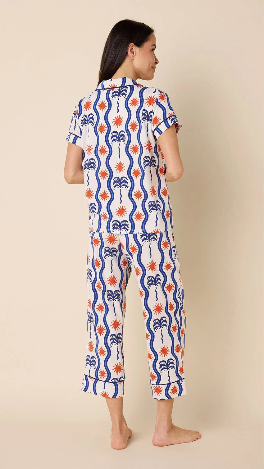 A person is smiling while wearing the luxurious CAT&#39;S PAJAMAS Red, White and Breeze Pima Knit Capri Set. This stylish loungewear features a short-sleeve button-up top with blue and red abstract designs on white fabric, paired with chic cropped pants, set against a beige background.