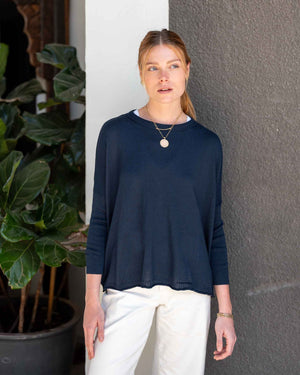 A woman stands in front of a gray wall and a green potted plant, wearing the MERSEA - CATALINA CREWNECK SWEATER IN NAVY by MERSEA with an oversized fit, paired with white pants, layered necklaces, and a bracelet, as she looks slightly to the side.