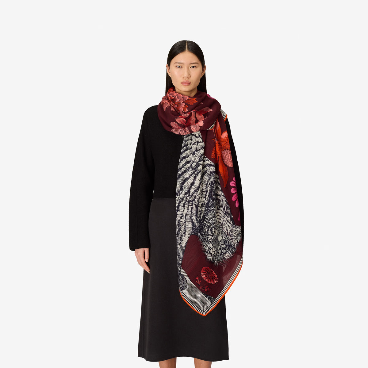 A woman with straight dark hair wears a black sweater and the INOUI EDITIONS - GUSTAV WOOL SCARF IN BURGUNDY by INOUI—a large (130 x 130 cm), multicolored patterned scarf crafted in India with cozy wool in rich red, pink, and burgundy tones—set against a plain white background.