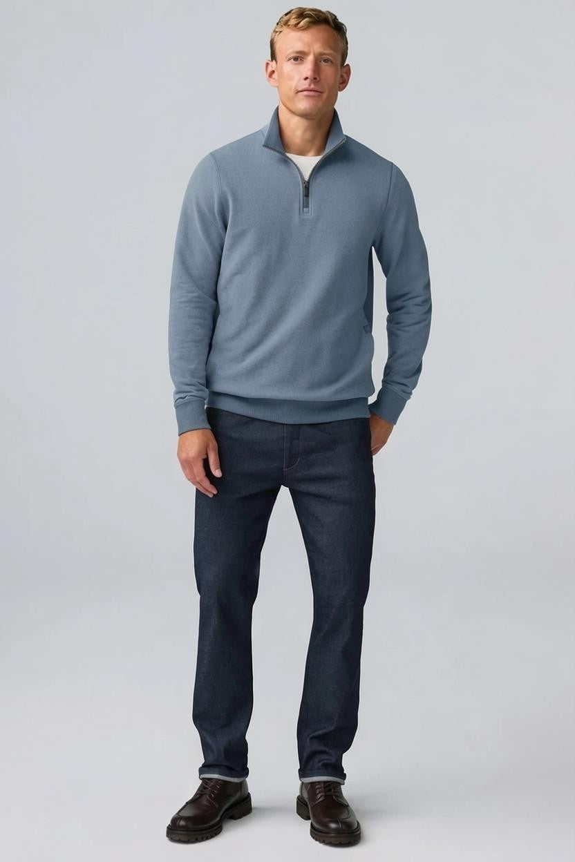 A man with short light brown hair stands in profile, wearing the MACK WELDON Ace 1/2 Zip Sweater in Herringbone and dark pants, one hand in his pocket, against a plain light gray background.