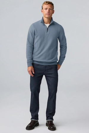 A man with short light brown hair stands in profile, wearing the MACK WELDON Ace 1/2 Zip Sweater in Herringbone and dark pants, one hand in his pocket, against a plain light gray background.