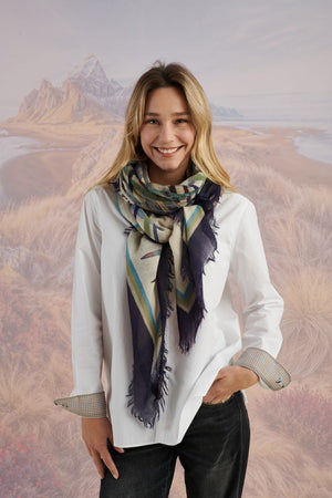 A woman in a blue shirt and jeans stands holding the STORIATIPIC - JULIE WOOL SCARF, a luxury plaid accessory in earthy tones. Behind her, mountains, beach, and grassy dunes create a scenic backdrop.