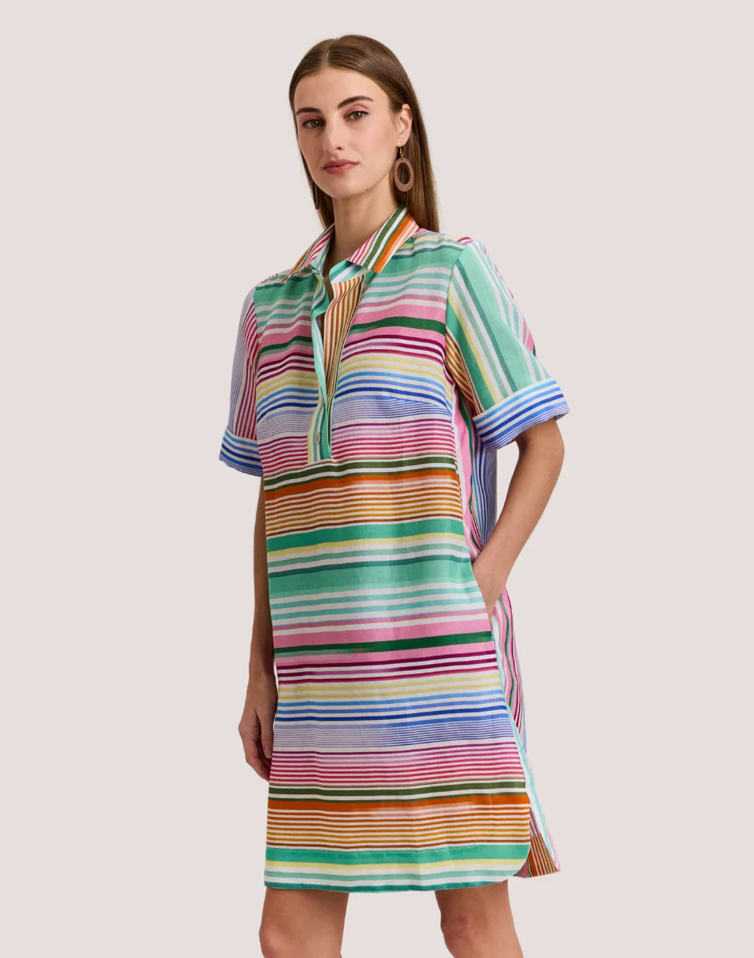 A woman wears the HINSON WU AILEEN Short Sleeve Linen Dress in Multi Stripe, a knee-length design with colorful stripes, a collar, front buttons, and pockets.