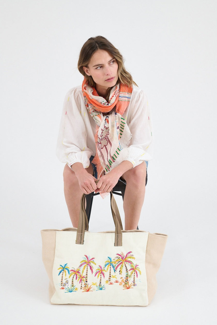 A person in a white shirt and blue jeans holds the STORIATIPIC BEACH TOTE BAIA, an embroidered cotton bag featuring colorful palm trees and plants.