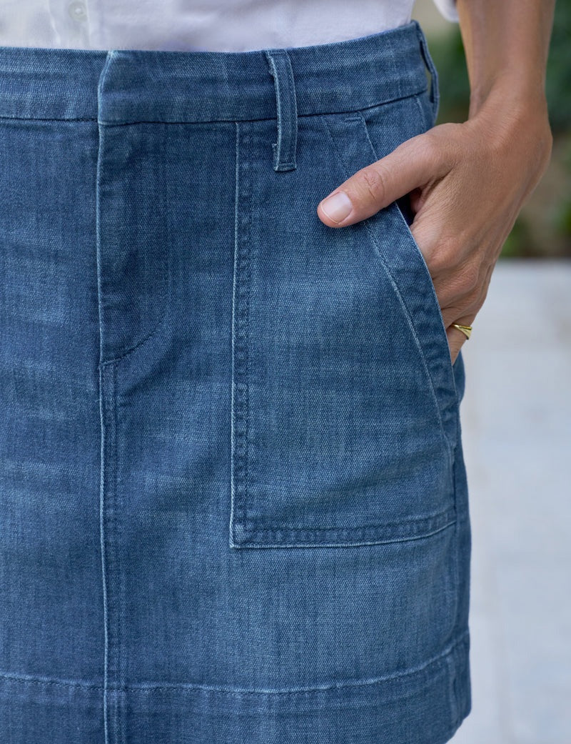 Close-up of a person wearing the FRANK &amp; EILEEN Jameson Utility Skirt in Italian Dream Light Denim 1984 Wash, with utility pockets, a white shirt, and a gold ring. Their right hand rests in the front pocket; background is blurred.