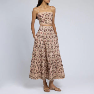 A woman is wearing a light pink floral two-piece outfit. The SABINA SAVAGE - HONOURING ARGOS CROPPED FITTED TOP from SABINA SAVAGE ENGLAND is sleeveless with thin straps and a square neckline, paired with a high-waisted skirt that flows gracefully from the hips. Crafted from pure linen, the fabric features a pattern of small, detailed floral designs perfect for any Greek-inspired look.