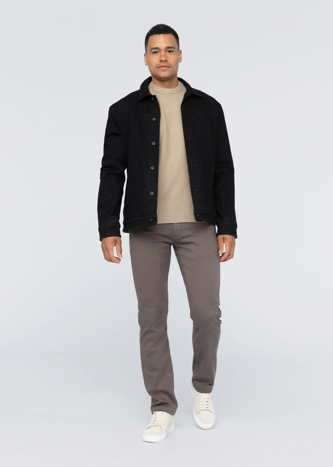A person faces forward against a plain light background, wearing a black jacket over a beige sweater, DUER No Sweat Straight Pants in Canteen, and white sneakers.