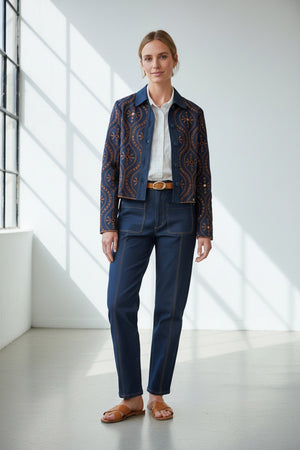 A woman stands indoors by large windows, wearing the MERSEA - FIFI JACKET—an indigo cotton jacket with cutwork embroidery—over a white shirt, paired with belted high-waisted blue pants and brown sandals. Sunlight creates grid-like shadows on the floor and wall.